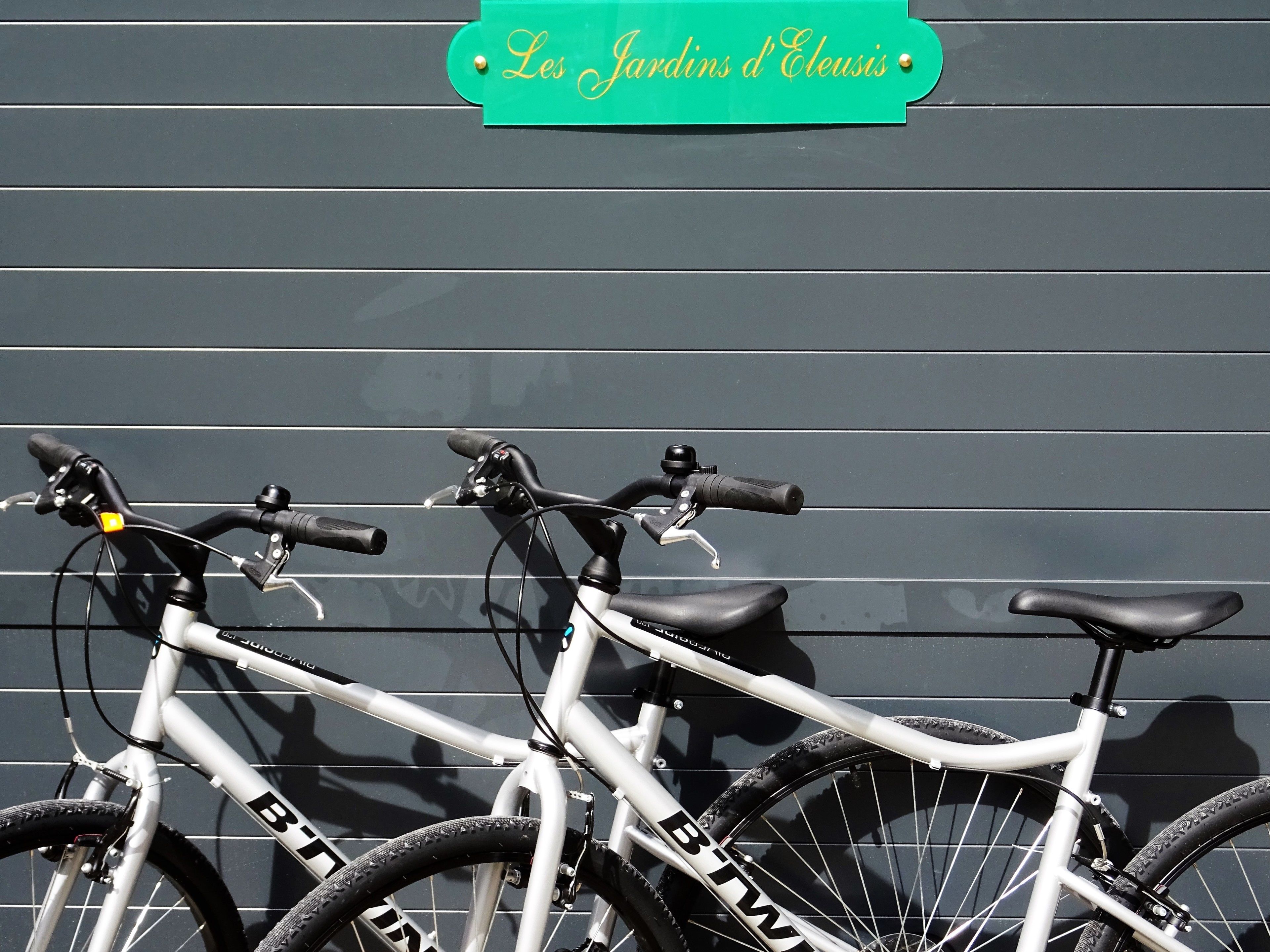 Two bicycles parked against a wall with a sign above them that reads "Les Jardins d'Eleusis."