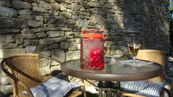 Un mur de pierres, des chaises en osier, une table en verre, une carafe d’eau rouge et un verre contenant une boisson.