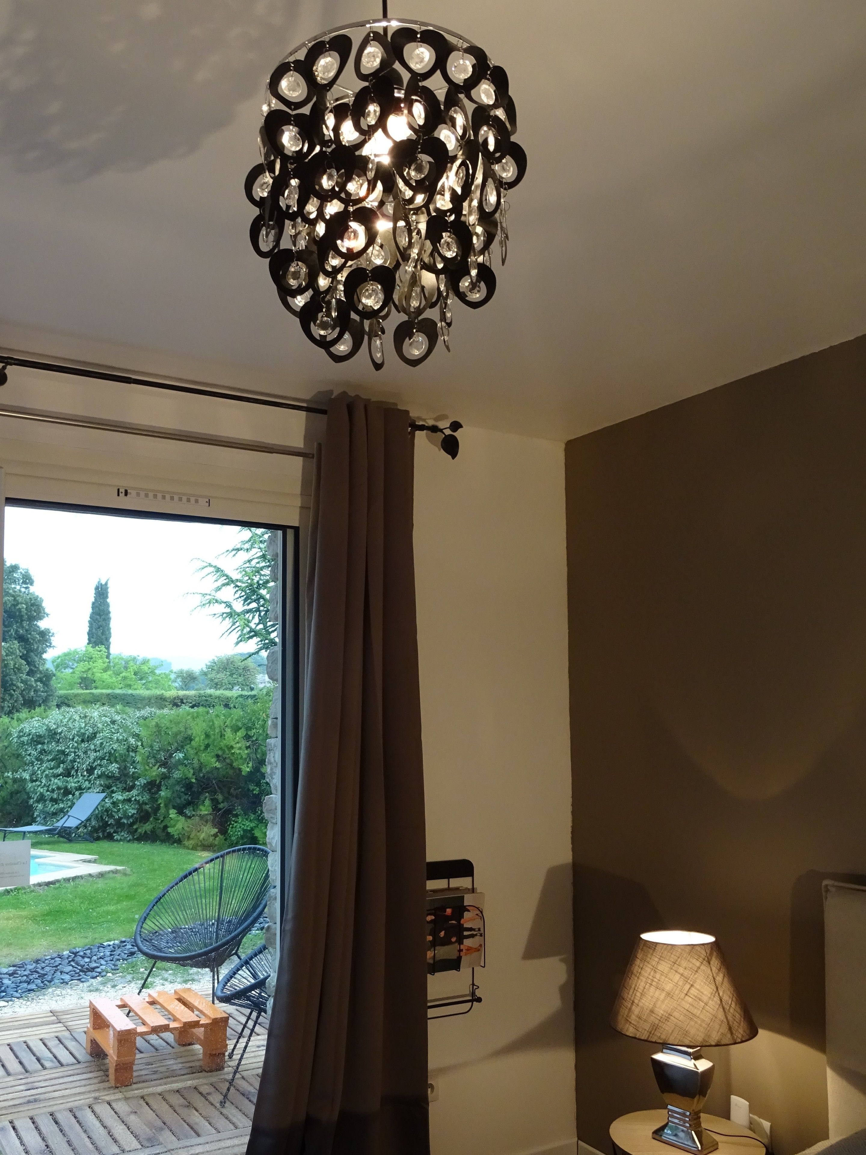 A room with a modern chandelier, a sliding glass door leading to a garden, a small wooden table, and a lamp on a bedside table.