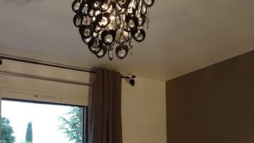 A room with a modern chandelier, a sliding glass door leading to a garden, a small wooden table, and a lamp on a bedside table.