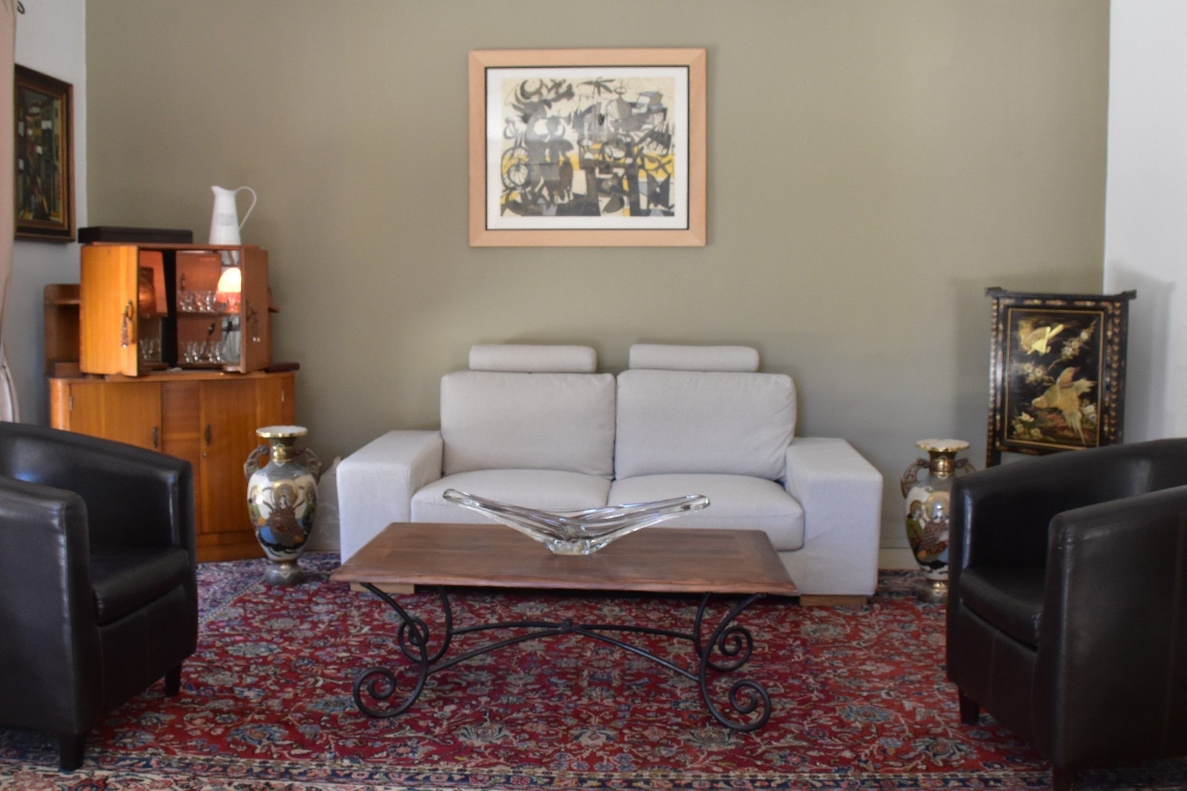 A living room with a white sofa, two black armchairs, a coffee table, a red patterned rug, and a framed abstract painting on the wall.