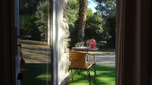 Vista a través de una puerta de vidrio que muestra una pared de piedra, una silla de mimbre y una mesa con un recipiente rojo y dos vasos.