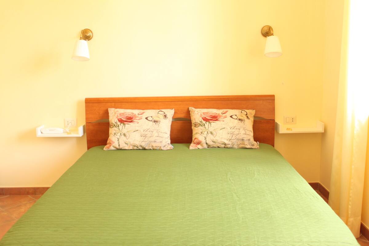 Een kamer met een groen sprei, twee bloemenkussens, een houten hoofdbord en twee nachtkastjes met lampen.