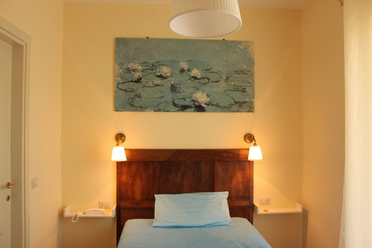 Een slaapkamer met een houten hoofdbord, een enkel bed met een blauw kussen, nachtlampjes en een schilderij van waterlelies aan de muur.