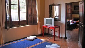 A bedroom with a bed, a television on a stand, a window with curtains, and a door leading to another room. - Casita La Chimenea (Antigua Guatemala)