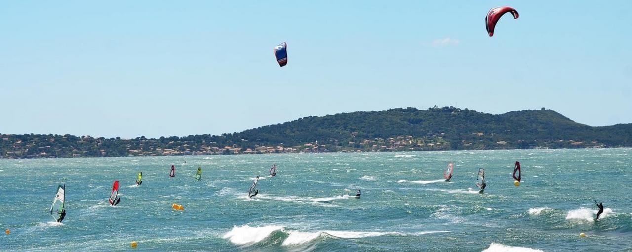 Een kustgezicht met meerdere windsurfers op het water, een heuvel in de verte met gebouwen, en een heldere lucht.