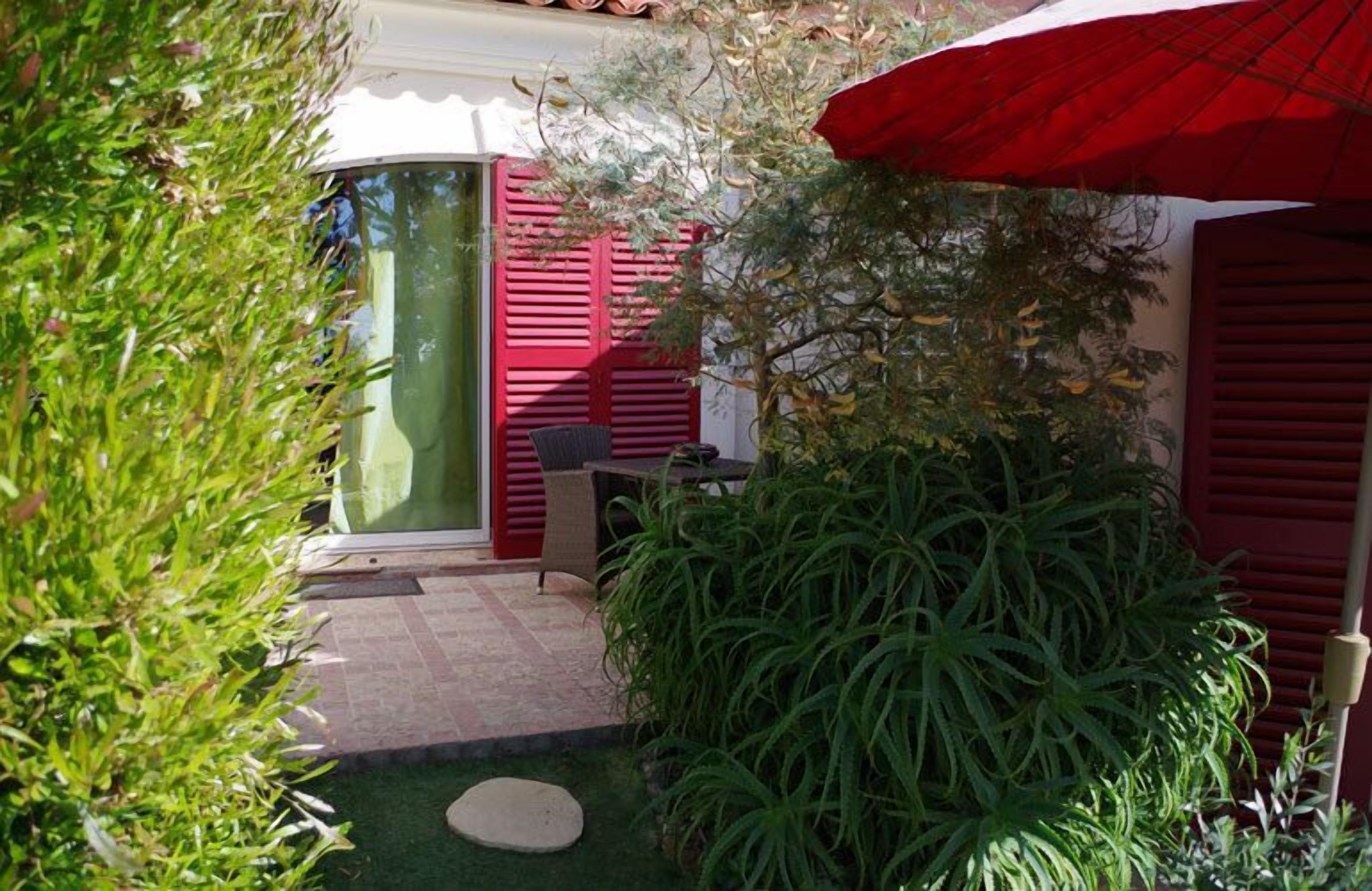 Un pequeño jardín vallado con una sombrilla roja, una silla de mimbre y una planta en maceta.