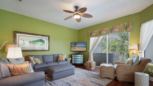 A living room with a sofa, armchairs, a coffee table, a TV, a ceiling fan, and a large window with curtains. - Sunkissed 3 Bedroom Townhouse by RedAwning (Holmes Beach)