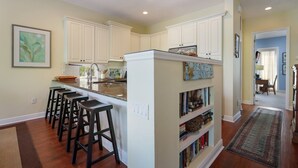A kitchen with white cabinets, a central island with bar stools, and a bookshelf. - Sunkissed 3 Bedroom Townhouse by RedAwning (Holmes Beach)