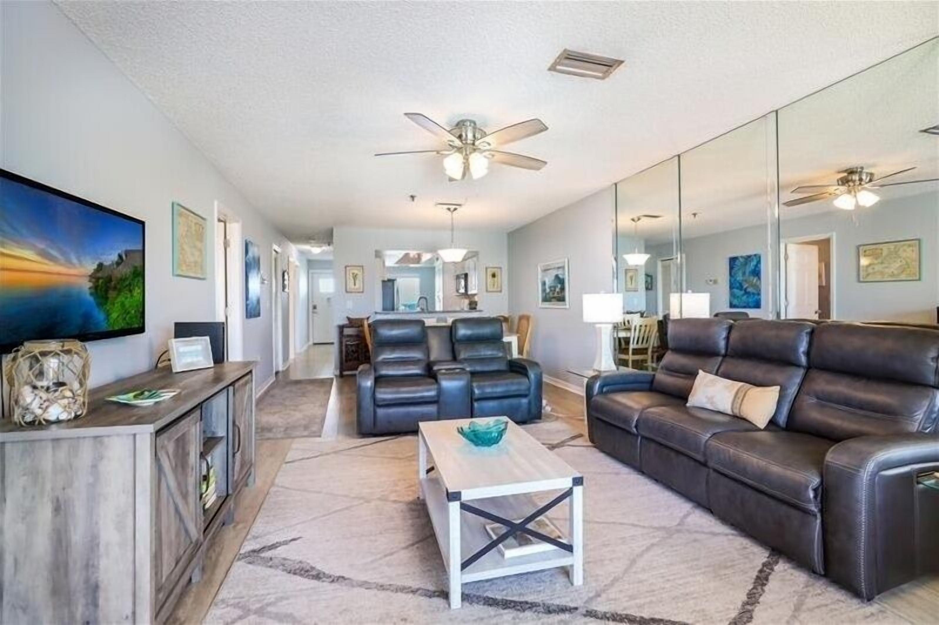A spacious living room with a large flat-screen TV, leather sofas, a coffee table, and a ceiling fan.