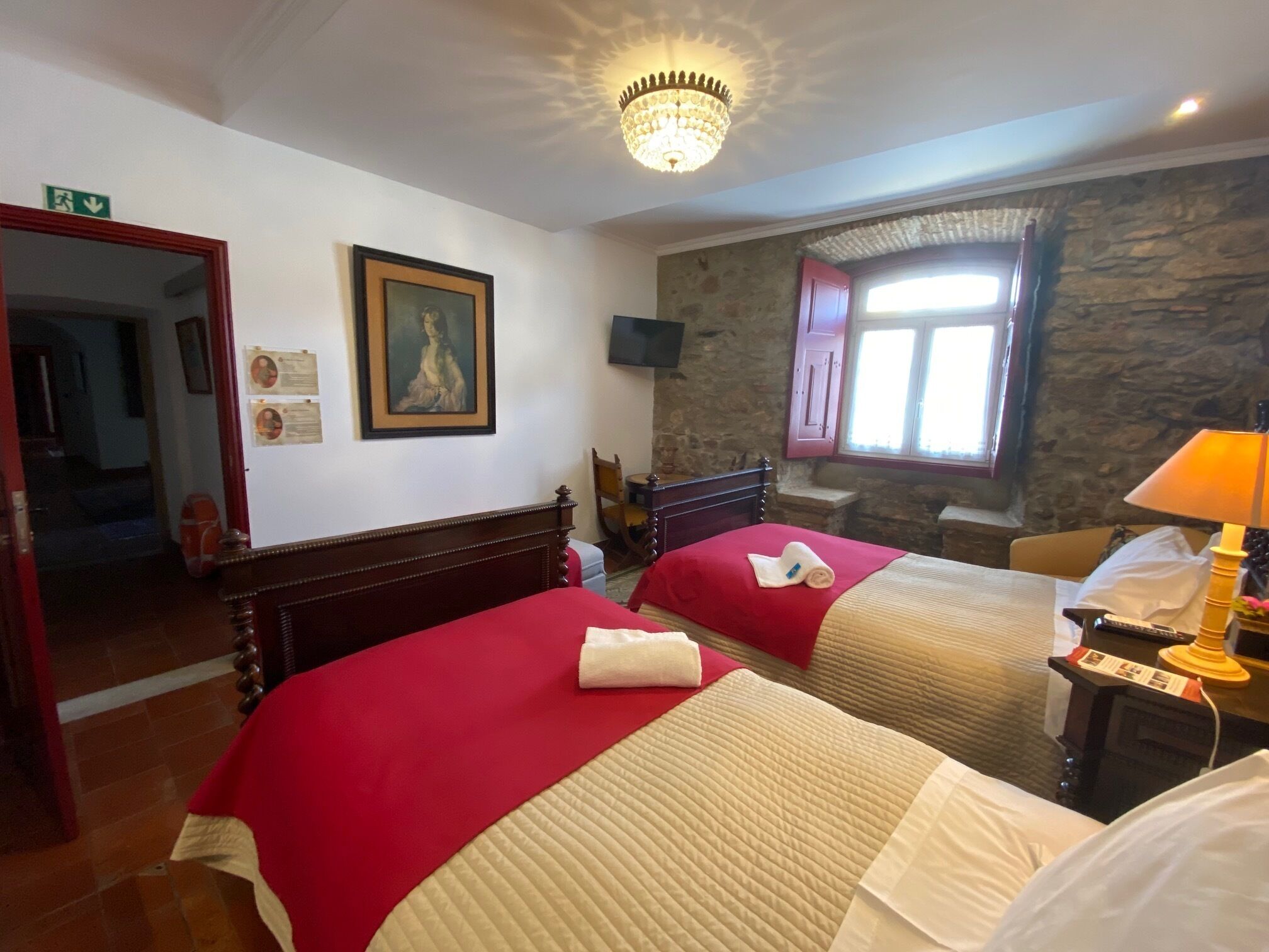 A hotel room with two beds, a chandelier, a stone wall, and a window with shutters.