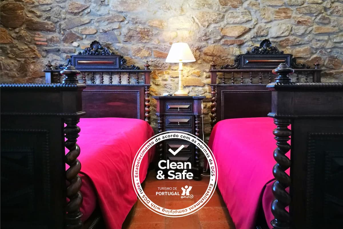 A room with a stone wall, two wooden beds with red bedspreads, a nightstand with a lamp, and a small table with a decorative item.