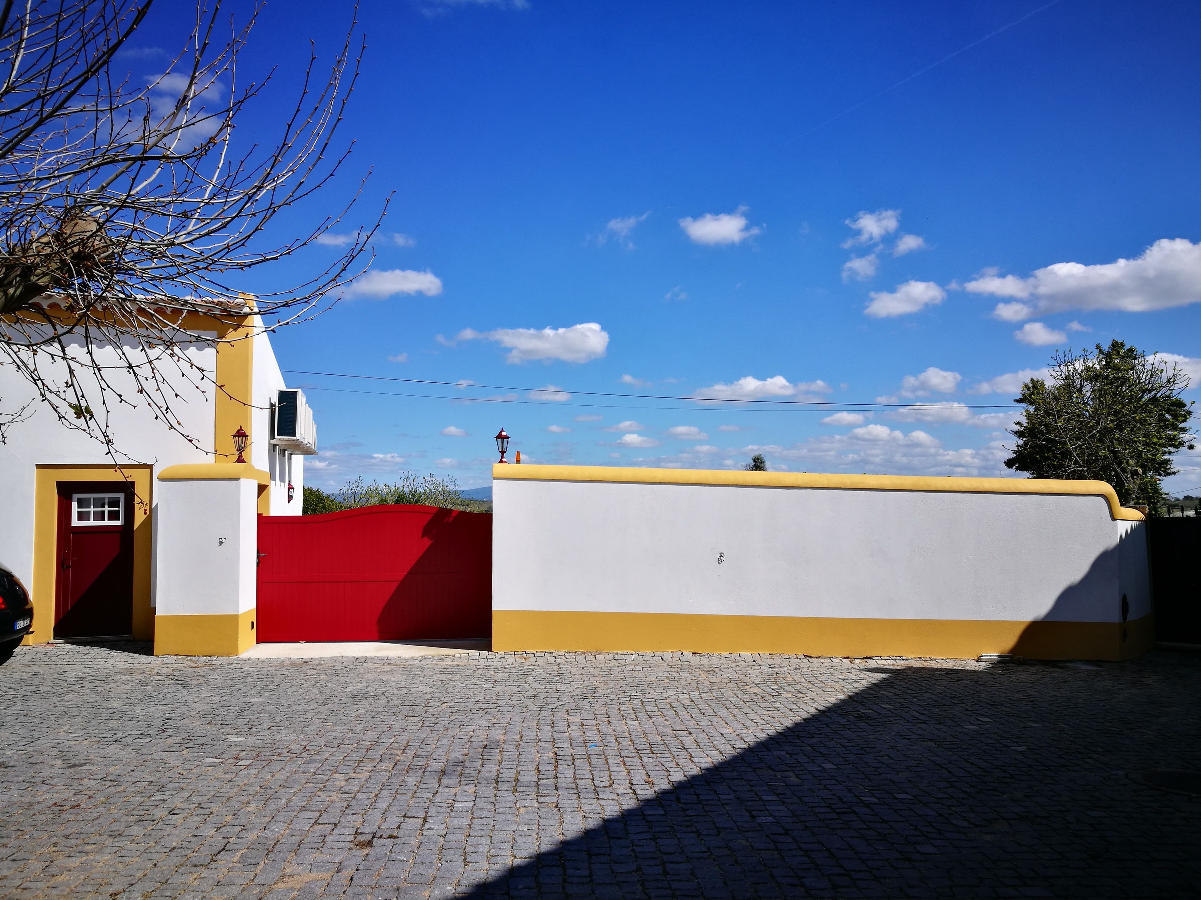 Un edificio con puerta roja y fachada amarilla y blanca, un patio de adoquines y un cielo azul despejado con nubes.