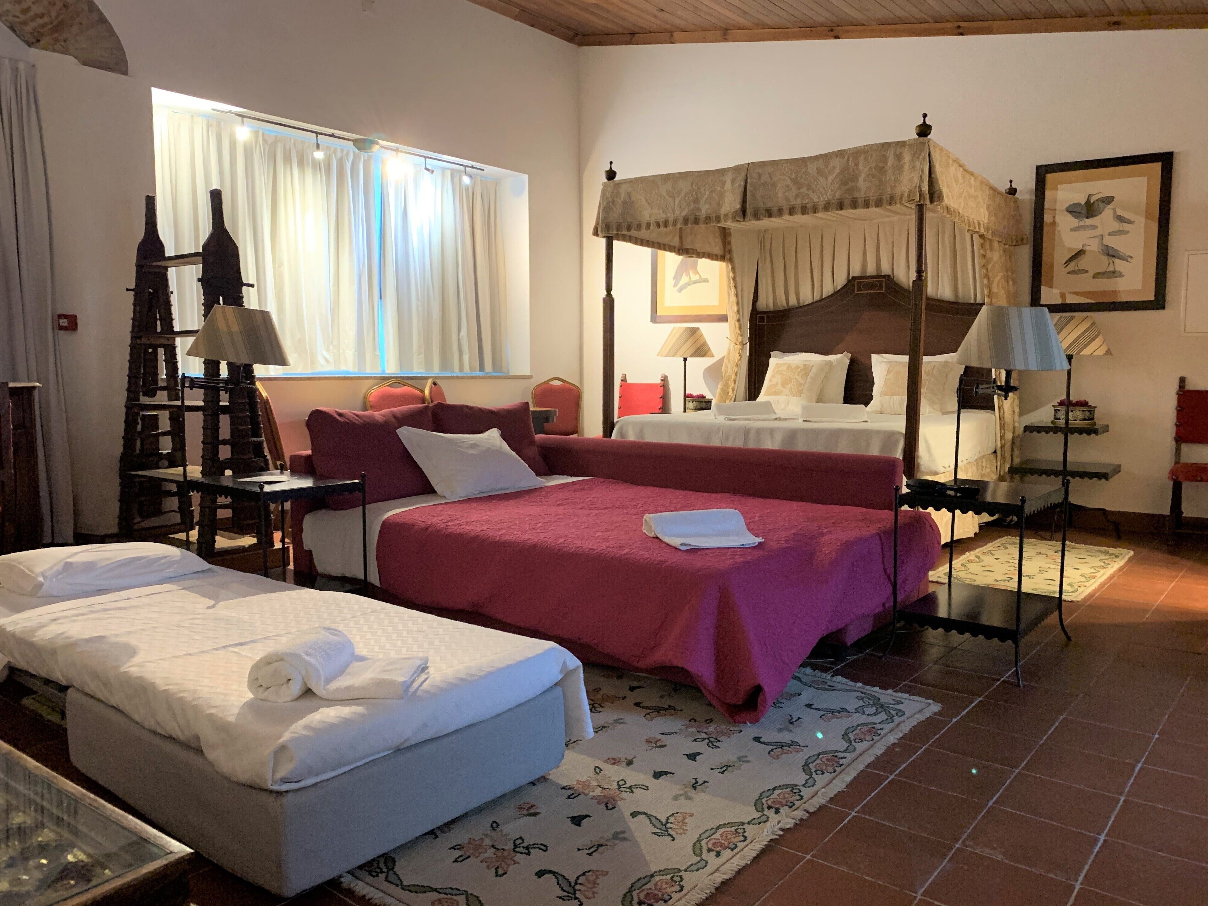 A hotel room with two beds, a wooden ceiling, a window with curtains, a bedside table with a lamp, and a framed picture on the wall.