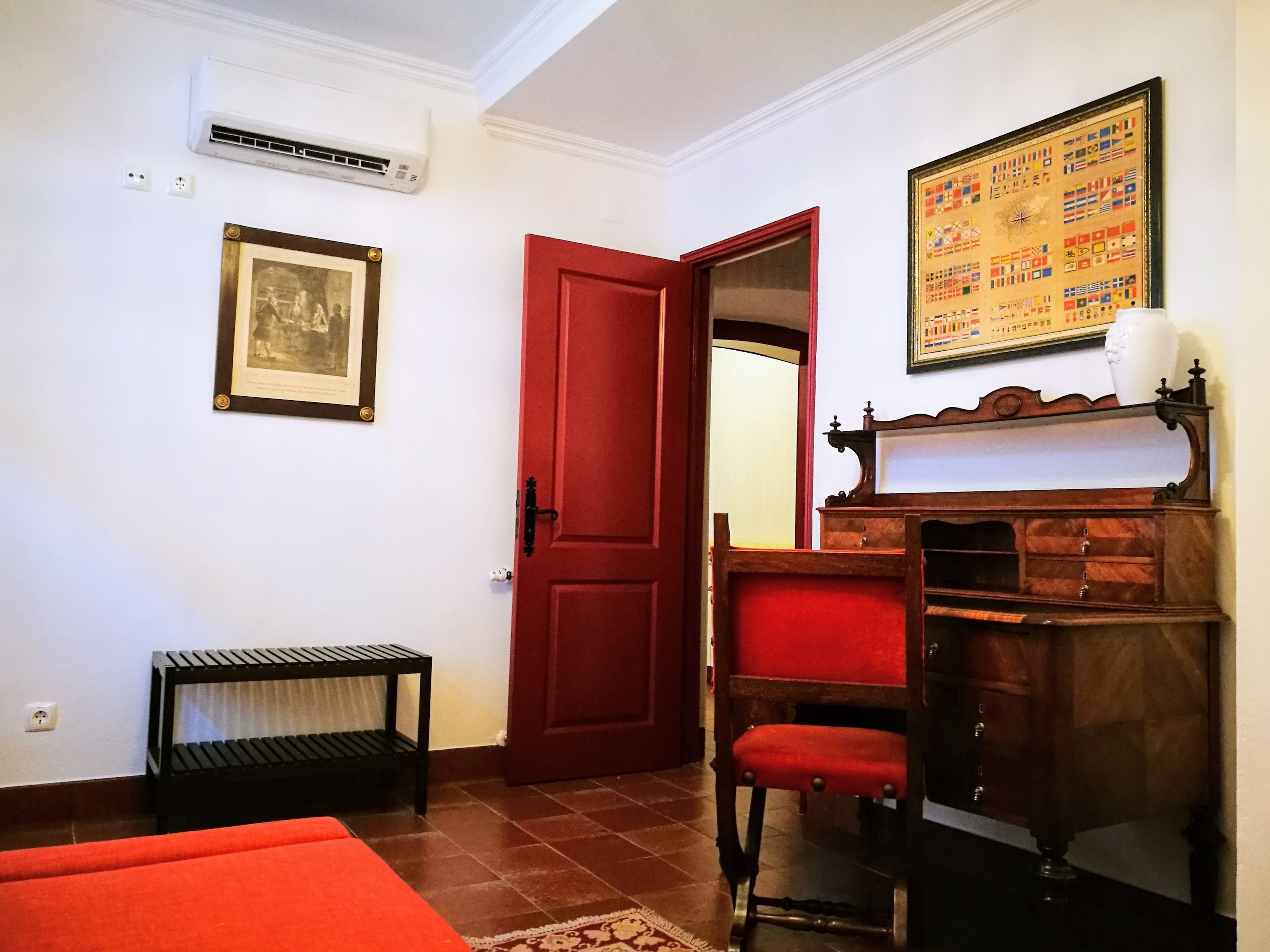 Una habitación con una puerta roja, un cuadro enmarco, un espejo, un armario de madera y una silla roja.