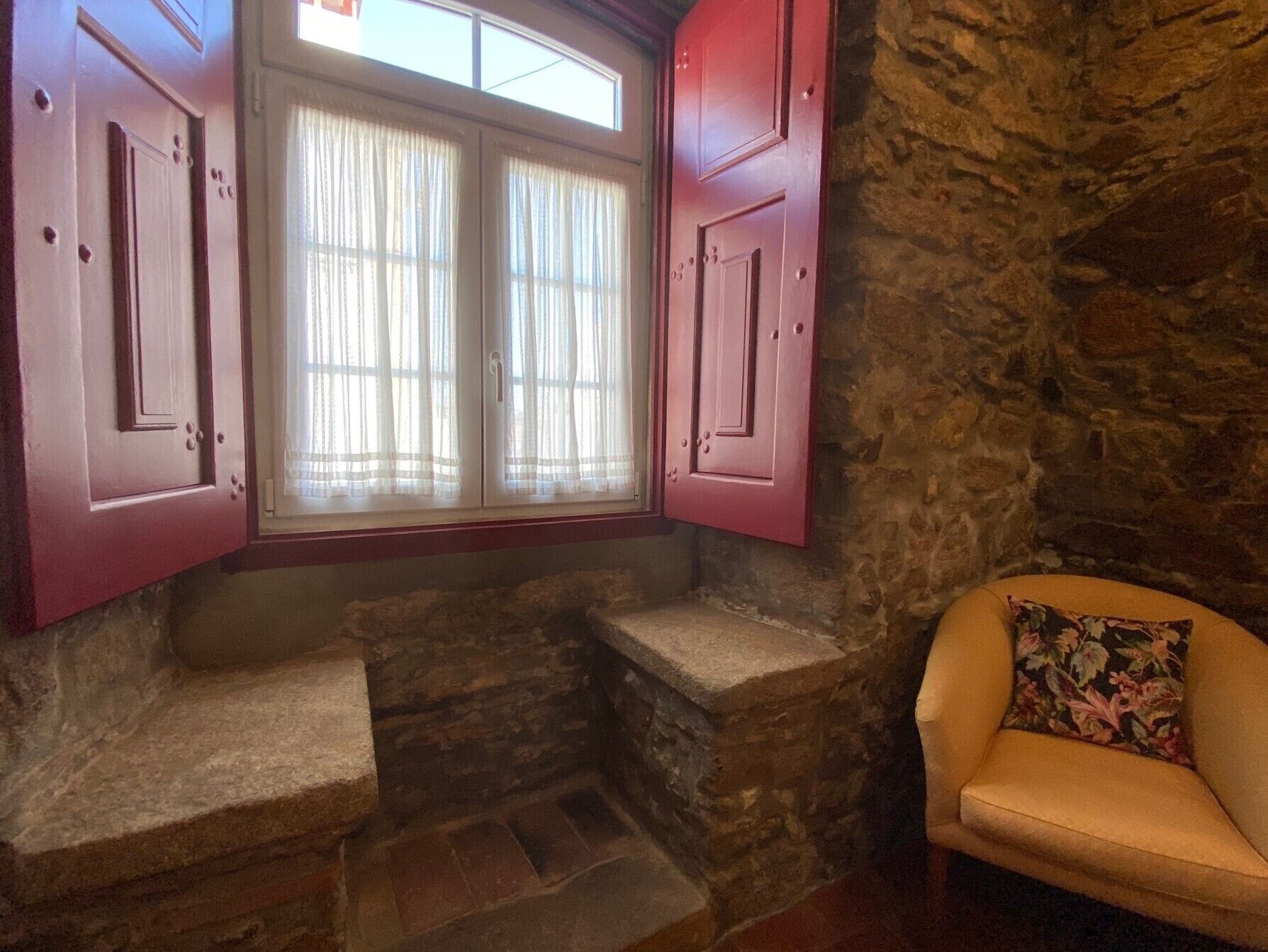 A stone-walled room with a window, a wooden bench, and a floral cushion.