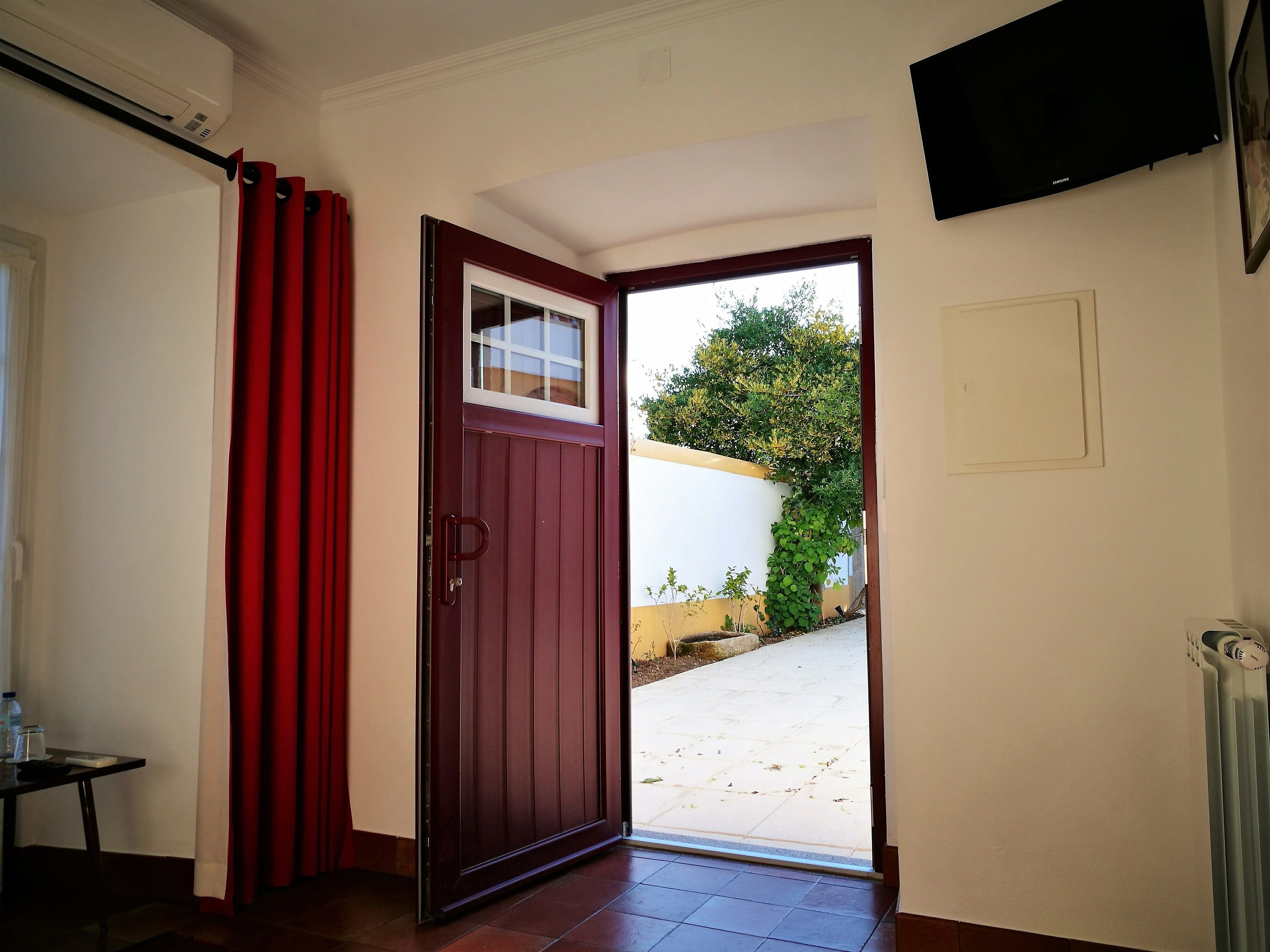 Una habitación con una puerta roja, una ventana, un radiador y un televisor fijado en la pared.