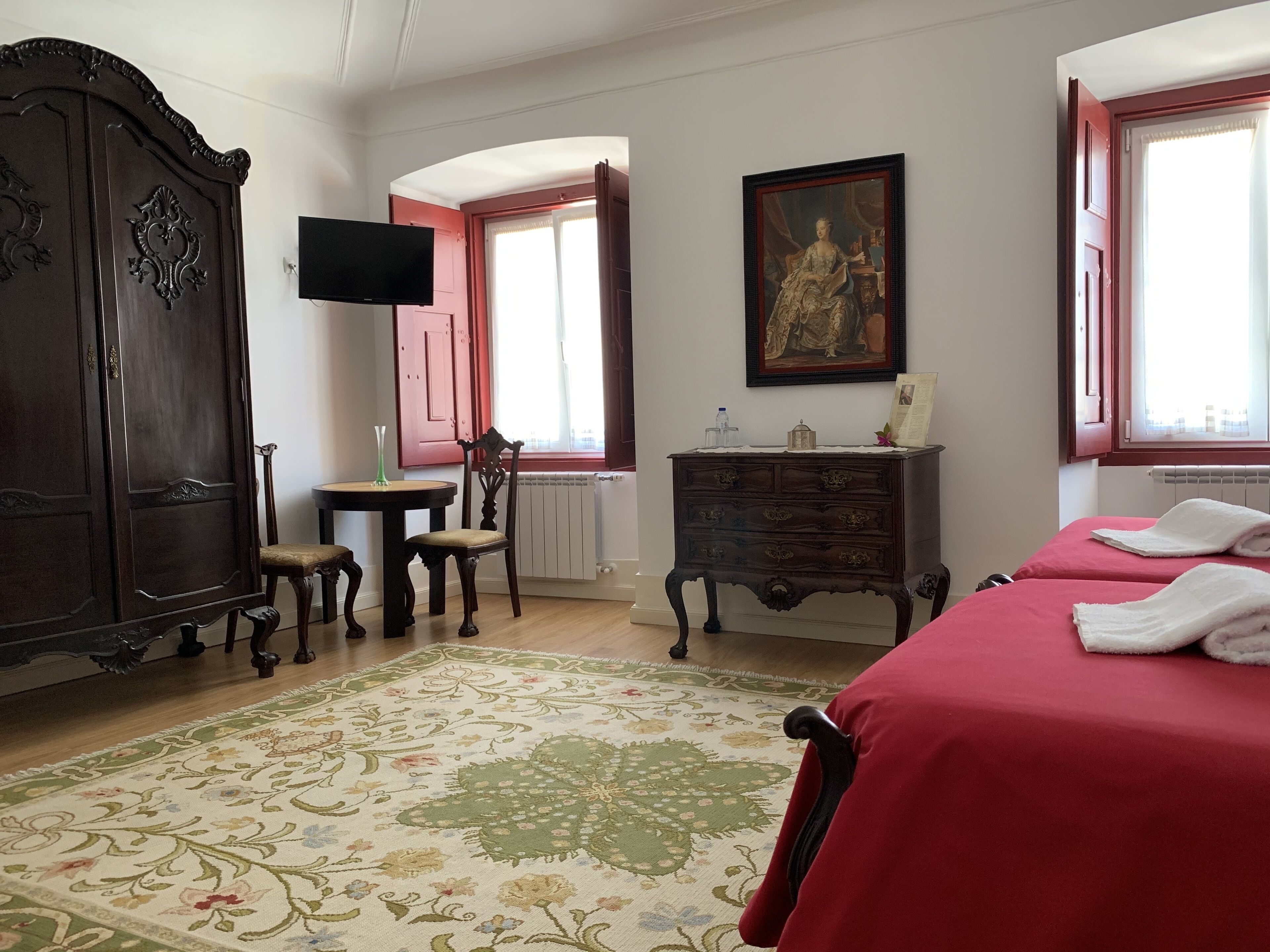 A room with a large wooden wardrobe, a small table with a vase, a chair, a dresser, a painting, and two beds with red bedding.