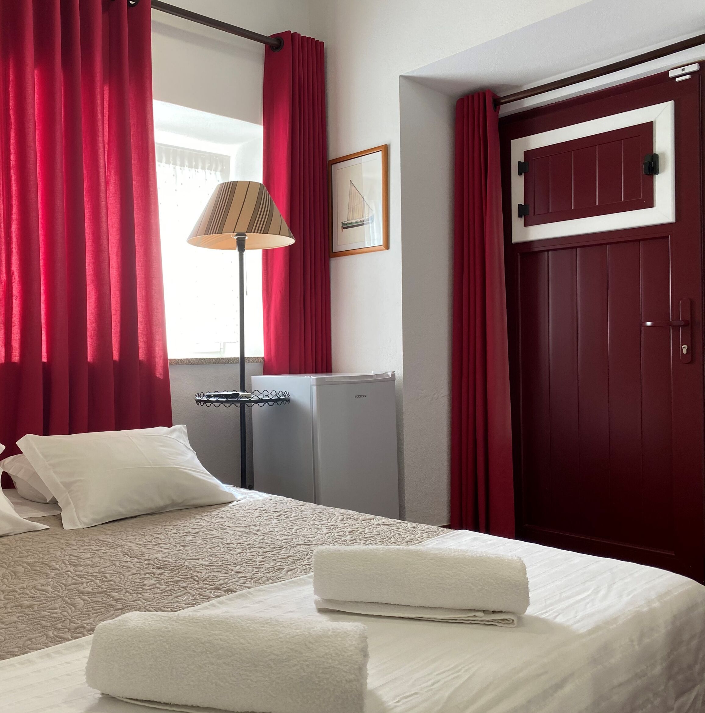 A hotel room with a bed, red curtains, a lamp, a framed picture, a door, and a small refrigerator.