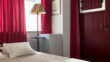 A hotel room with a bed, red curtains, a lamp, a framed picture, a door, and a small refrigerator.