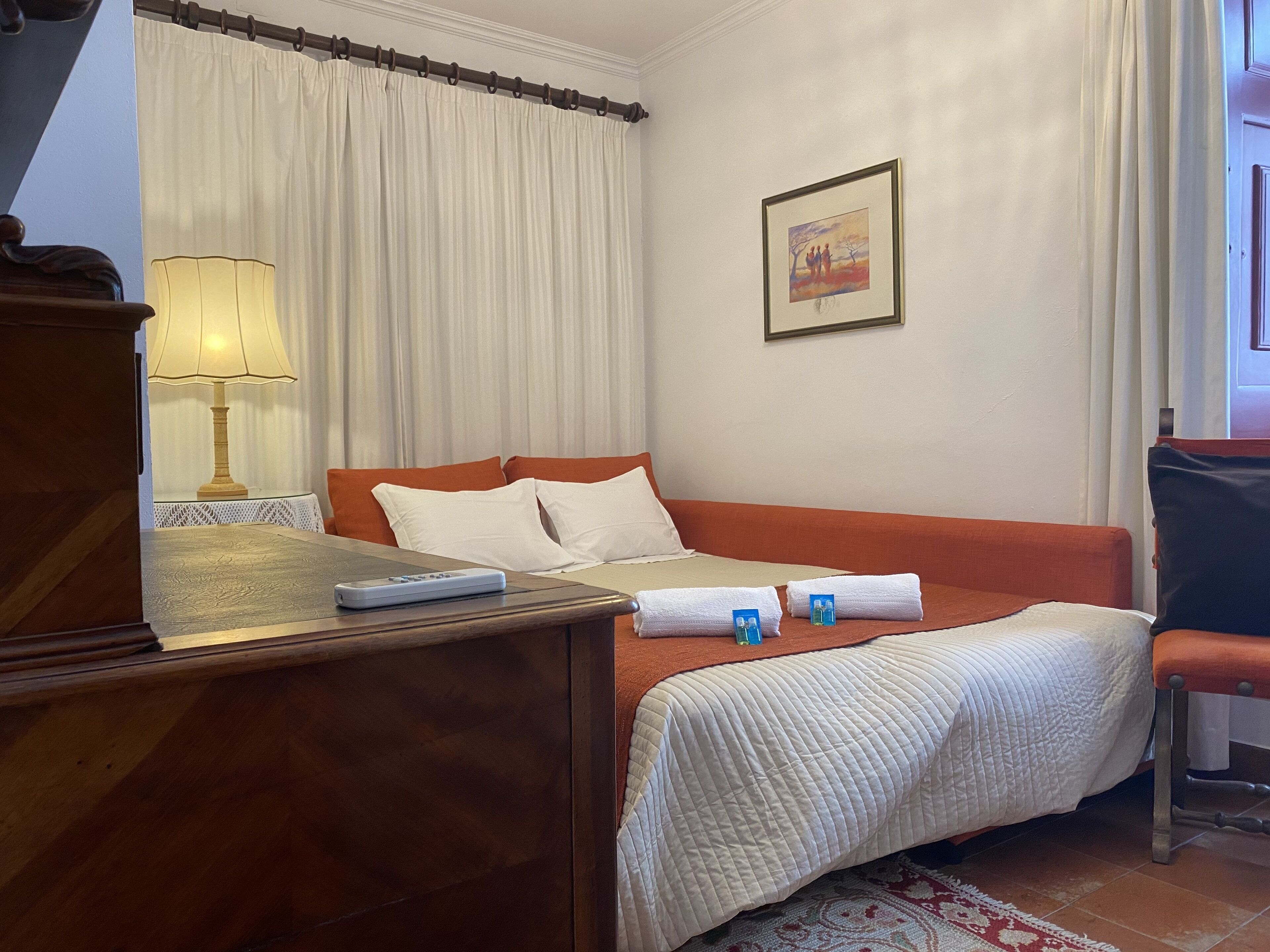 A hotel room with a bed, a bedside table with a lamp, a framed picture on the wall, and a window with curtains.
