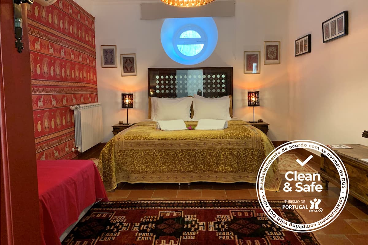 A hotel room with a bed, two bedside tables, a red patterned wall, a chandelier, and a circular window.