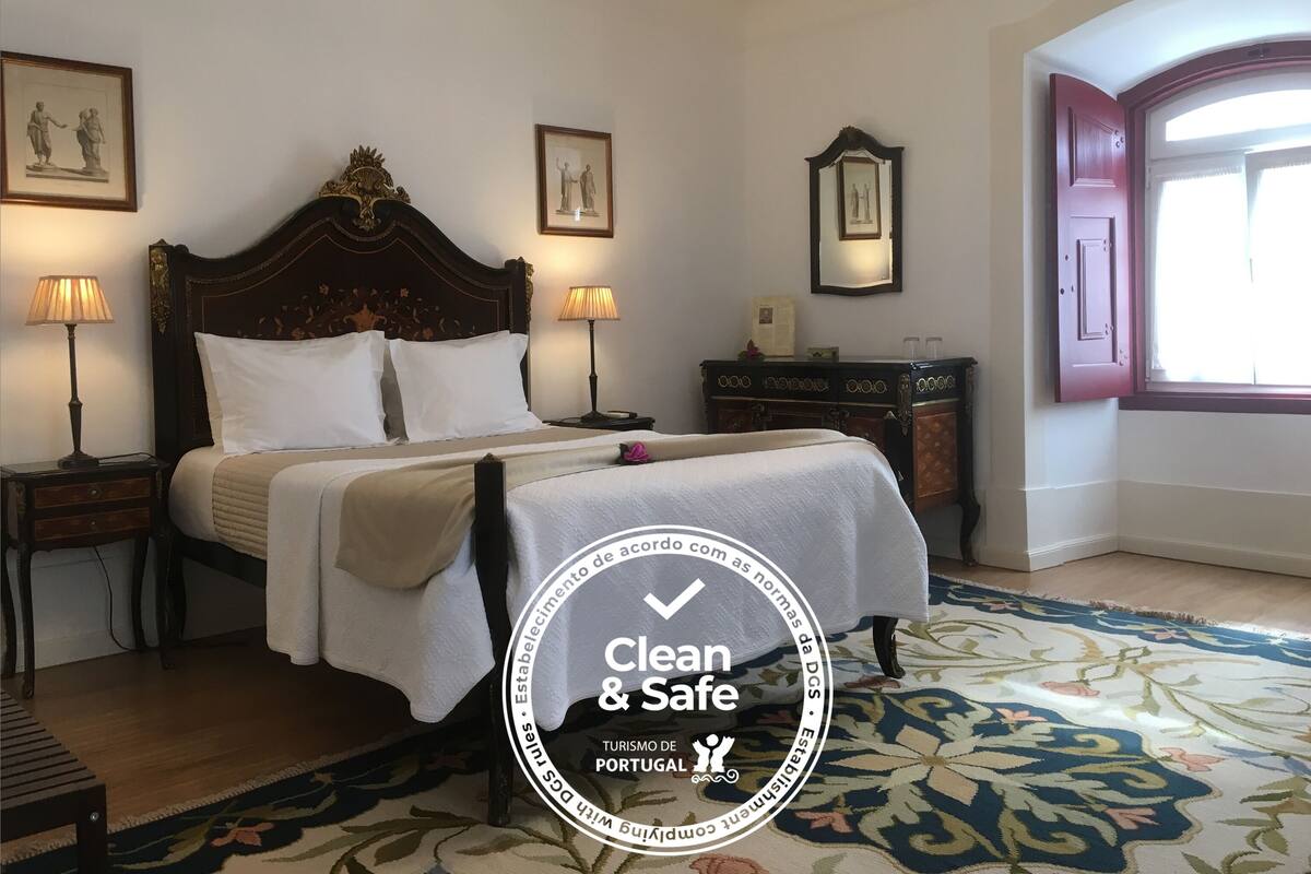 A hotel room with a bed, two bedside tables with lamps, a dresser, and a large window.