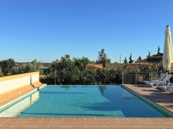 A clear blue pool with tiled edges, surrounded by lounge chairs and a white umbrella, set against a backdrop of trees and hills. - Casa do Peso (Crato)