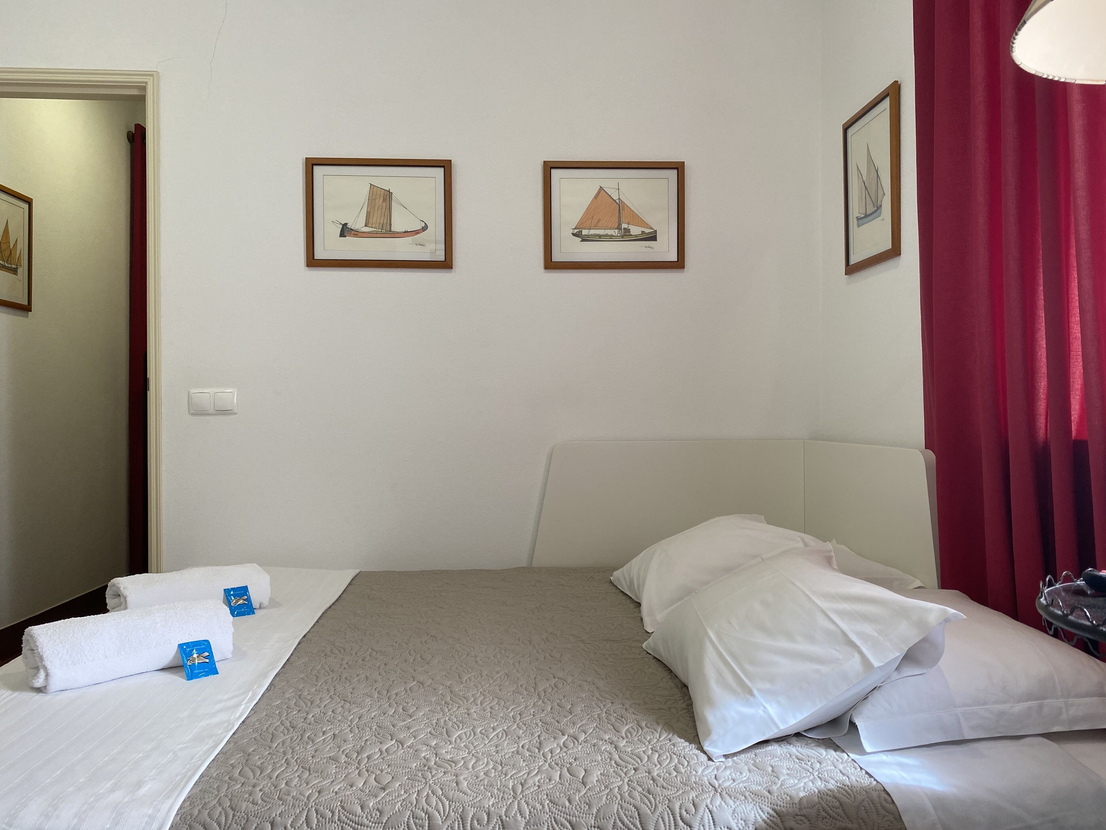 A hotel room with a bed, two framed pictures on the wall, a red curtain, and a white headboard.