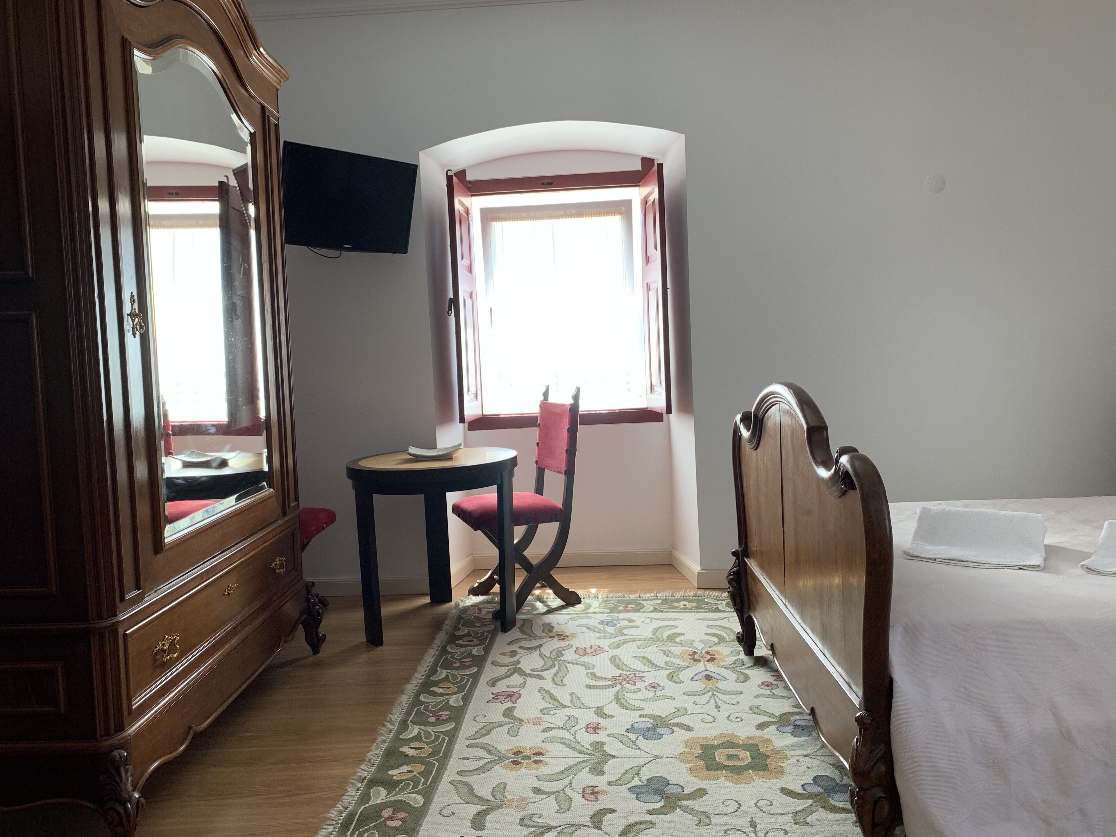 A room with a wooden wardrobe, a small table with a chair, a bed, and a television mounted on the wall.