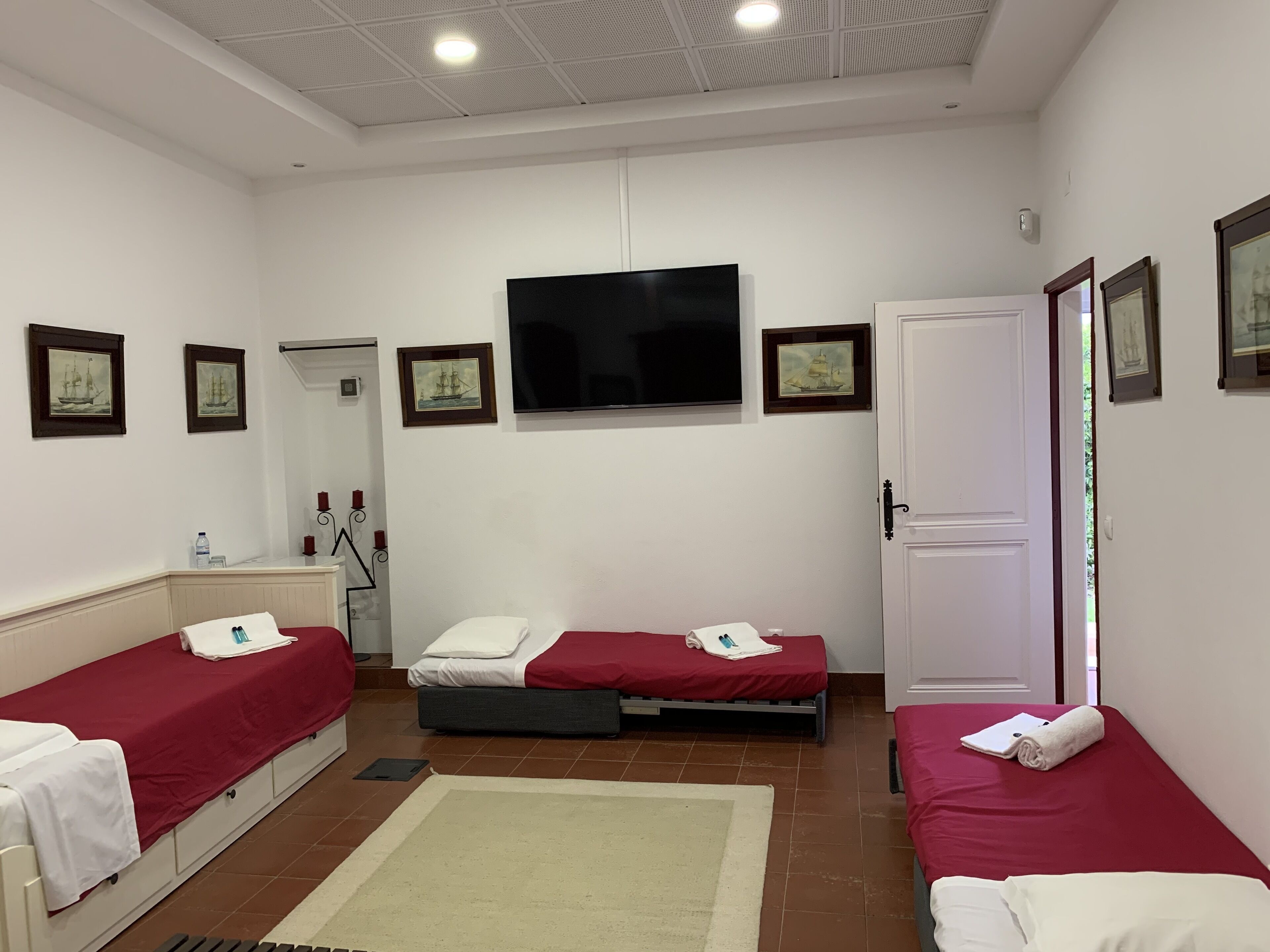 A hotel room with two single beds, a TV, a door, and framed pictures on the wall.