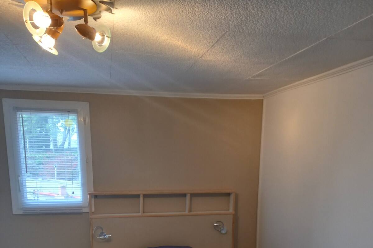A bedroom with a bed, bedside tables, a headboard, a window with blinds, and a ceiling light fixture.