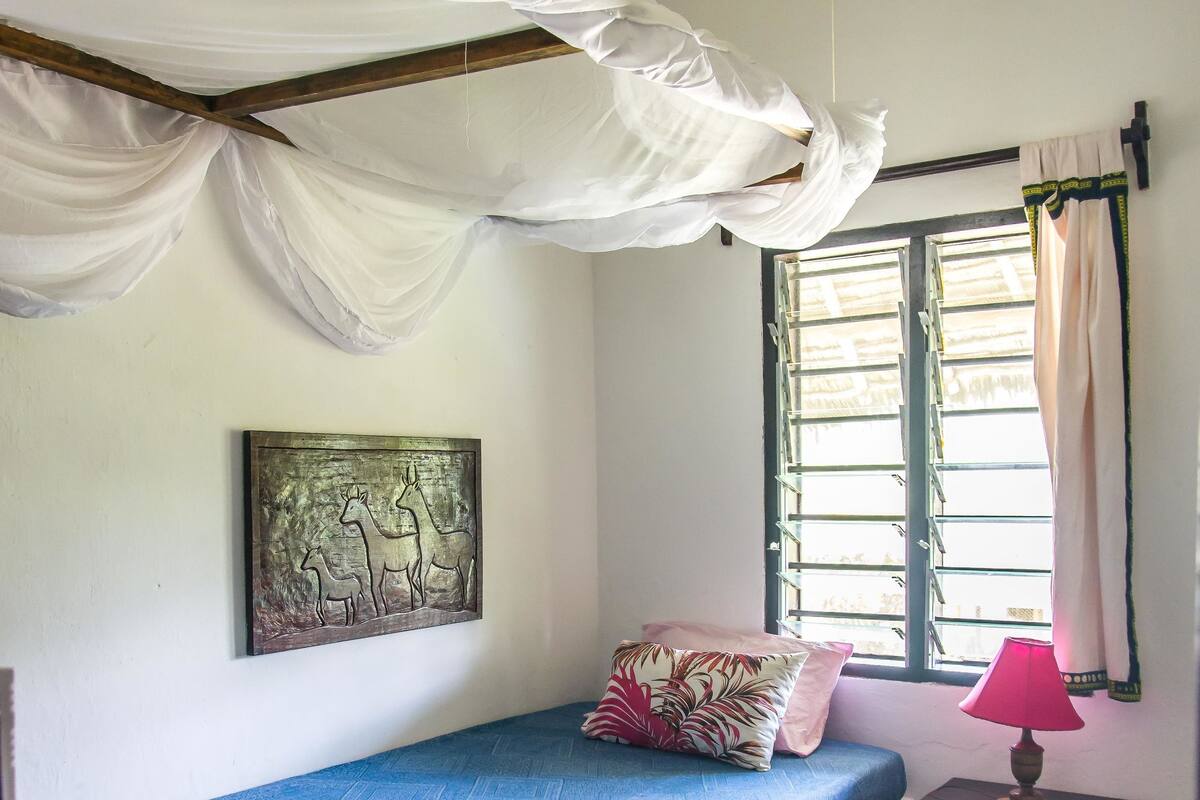A room with a bed, a small table with a lamp, a window with curtains, and a wall-mounted artwork.