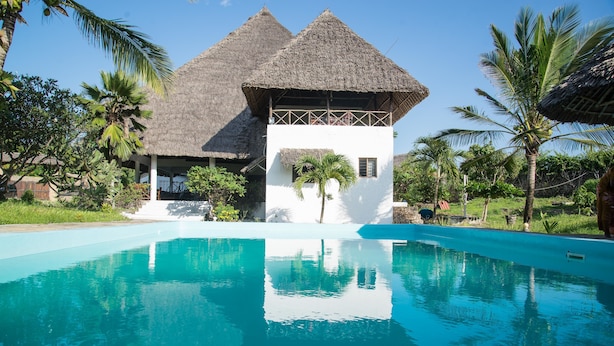 A poolside view of a resort with a thatched-roof building, a clear blue pool, and surrounding palm trees. - Ocean Breeze Villa (Watamu)