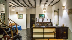 A room with exposed wooden beams, a wooden bench, a table with a blue tablecloth, and a bookshelf with various items. - Ocean Breeze Villa (Watamu)