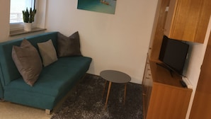 Una sala de estar con un sofá color aguamarina, una mesita redonda pequeña, un televisor sobre un mueble de madera, un cuadro de un lago y una ventana con persianas.