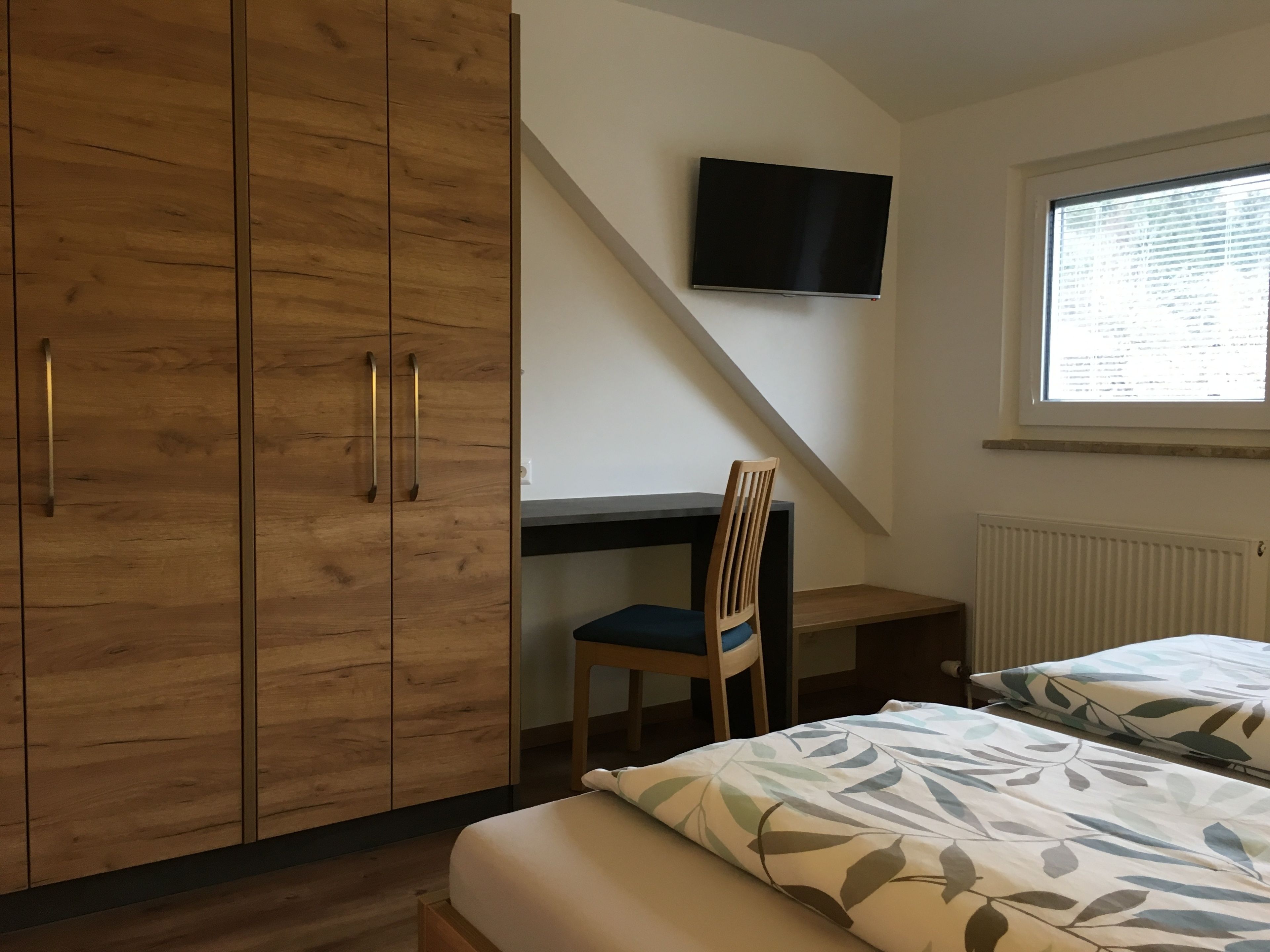 A bedroom with a desk, a chair, a bed with floral bedding, and a TV on the wall.
