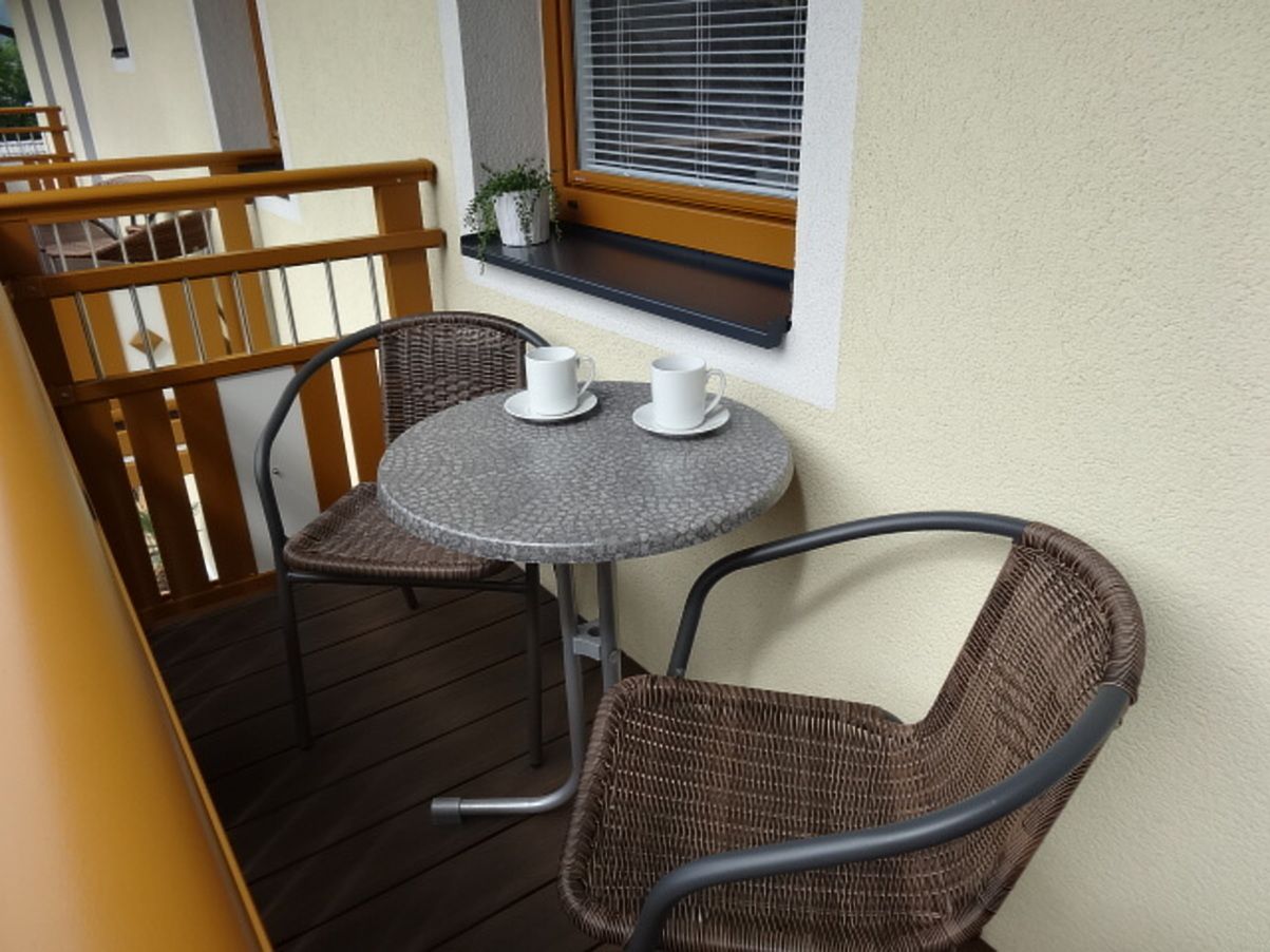 A small outdoor balcony with a round table and two wicker chairs, a window with blinds, and a wooden railing.