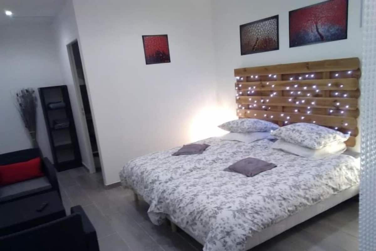 A bedroom with a bed, pillows, a headboard with lights, a red chair, and artwork on the wall.