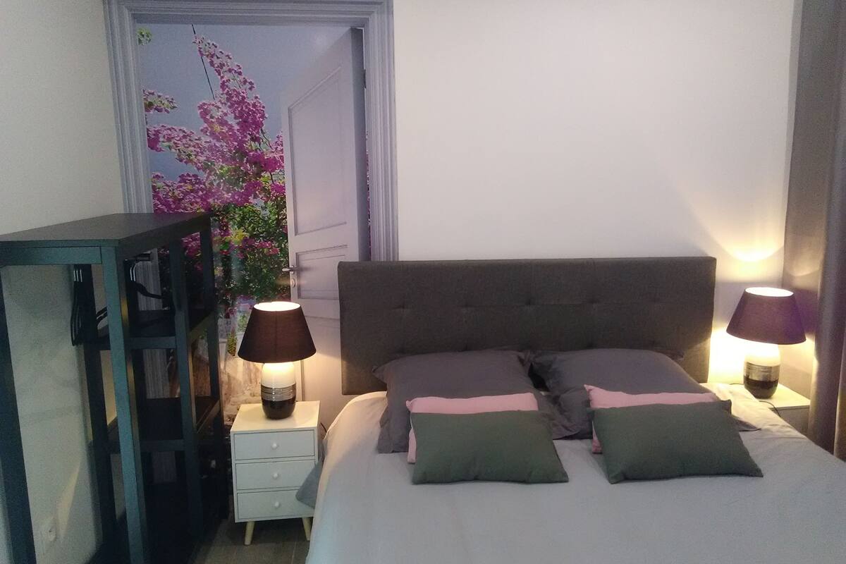 A bedroom with a bed, two bedside tables, a nightstand, a lamp, and a window with a view of flowers.