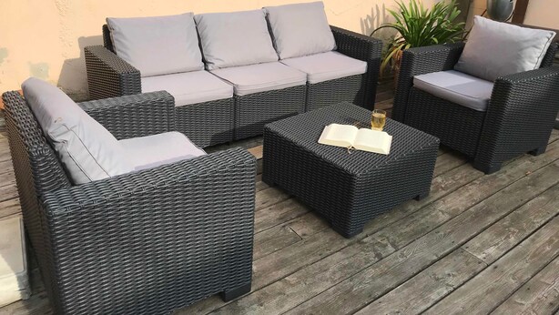 Un área de estar al aire libre con mimbre, cojines y una mesa, un libro y un vaso de bebida.