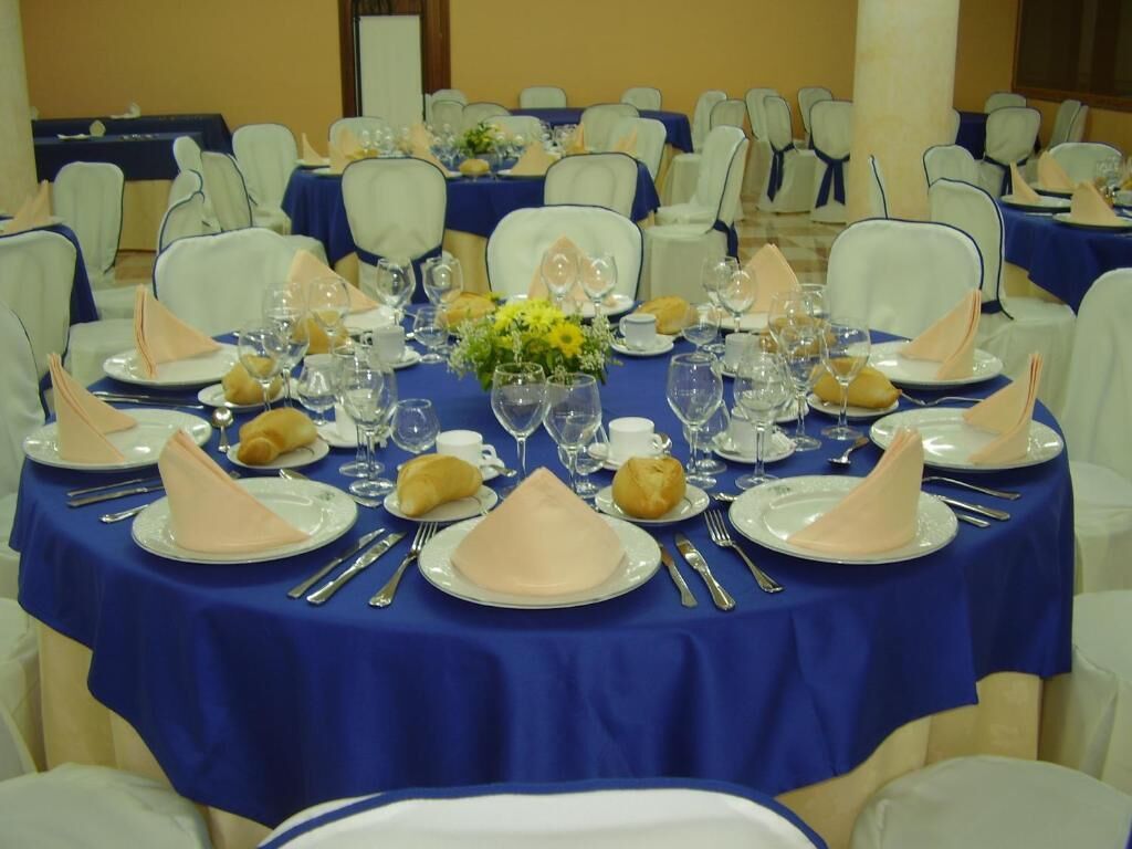 Un salón de banquetes con mesas redondas cubiertas con manteles azules, sillas blancas y cubertería ordenada.