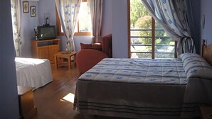Un dormitorio con una cama, un televisor sobre un mueble de madera, una ventana con cortinas y una mesita con una silla.