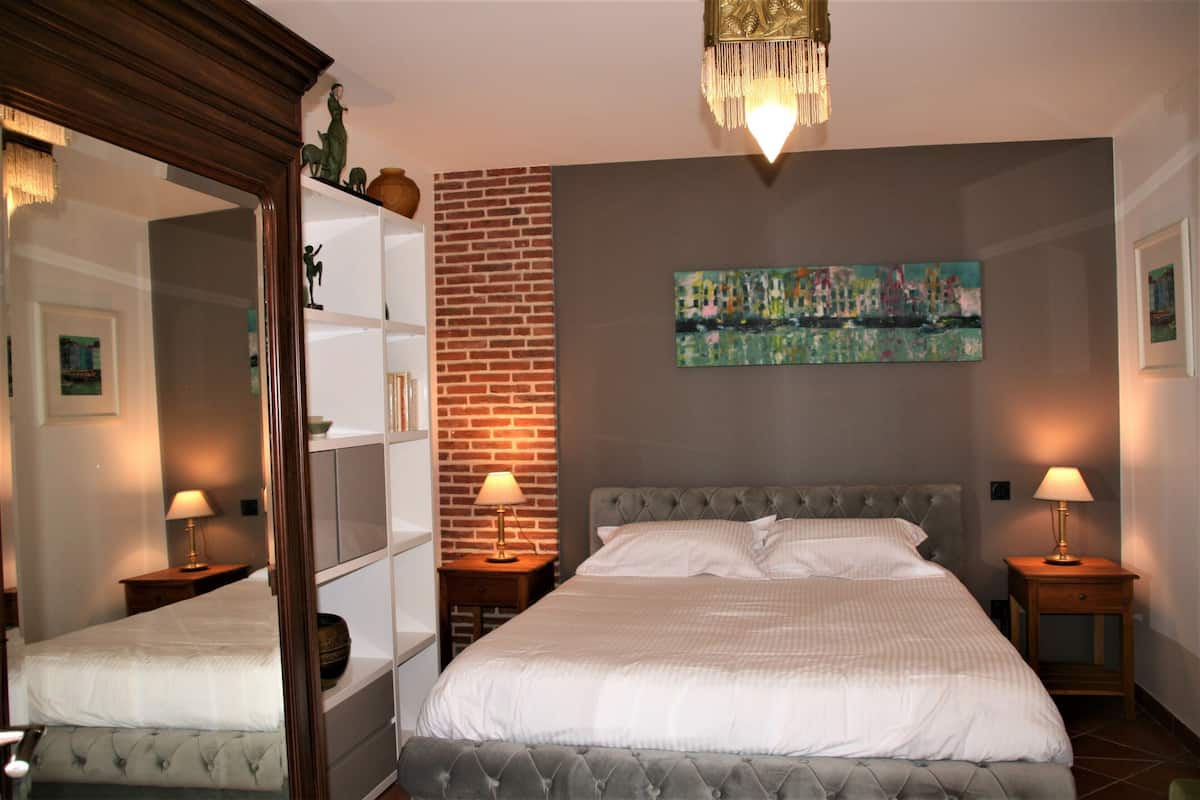 A bedroom with a large bed, a brick accent wall, a cityscape painting, and a chandelier.