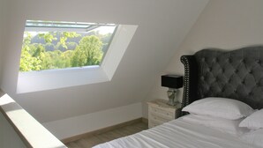 Un dormitorio con tragaluz, una cama con ropa de cama blanca, una mesita de noche con lámpara y vistas al exterior con vegetación.