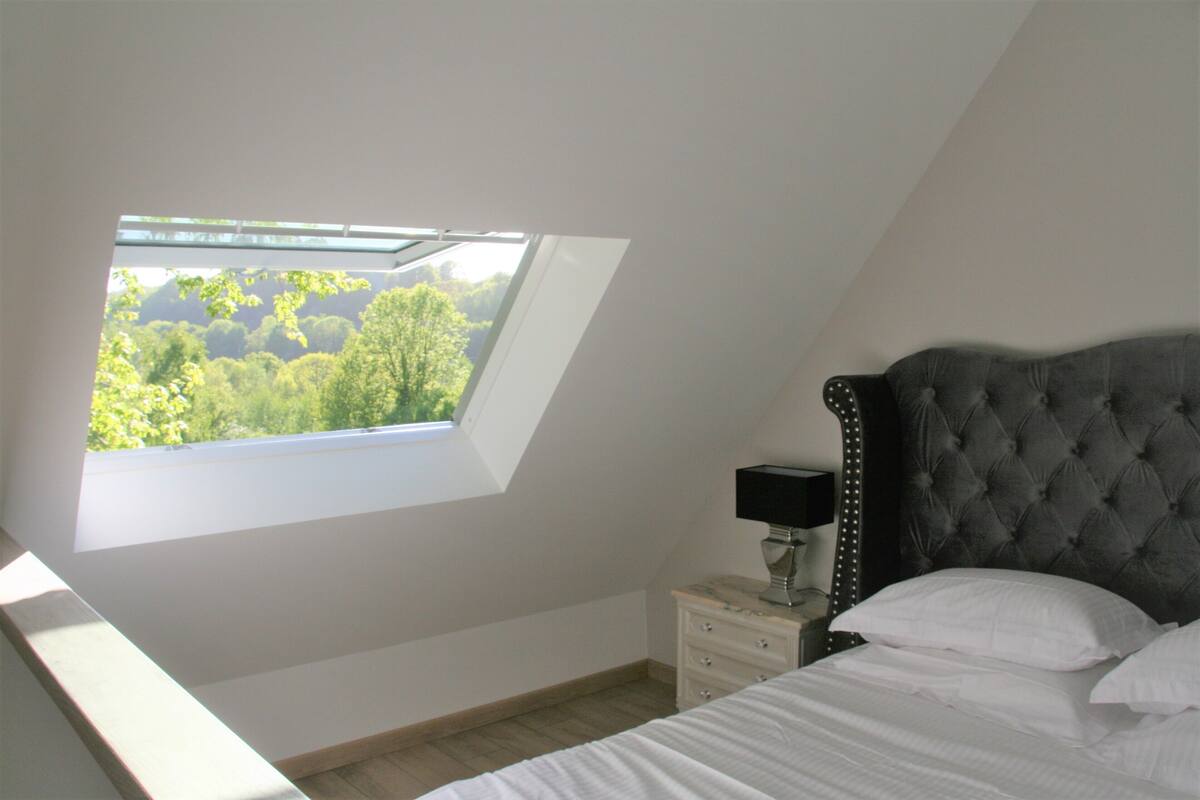 A bedroom with a skylight, a bed with white linens, a nightstand with a lamp, and a view of greenery outside.