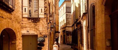 A narrow street with stone buildings, a woman walking, and a sign for 'Hotel Alchelejo'.