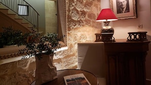 Una escalera que sube a un piso superior, una pared de piedra, una mesa con una lámpara, un retrato enmarcado y una planta en maceta.