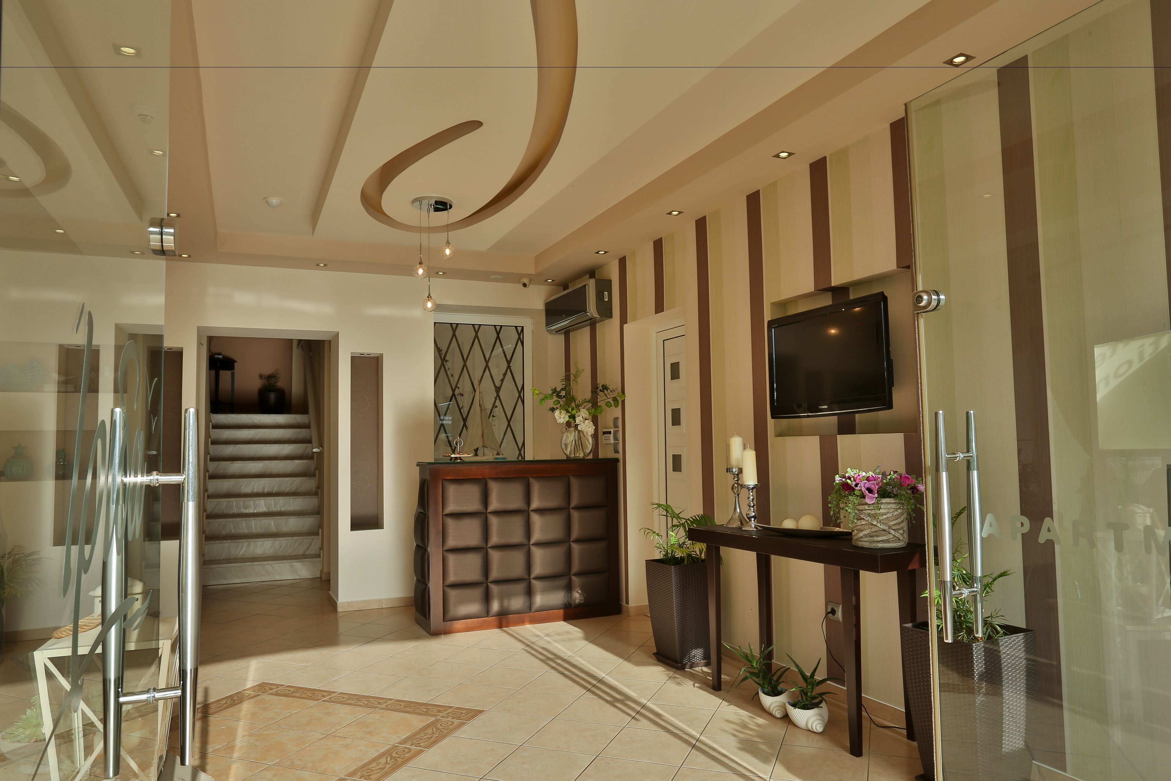 A modern hotel lobby with a reception desk, a staircase, and a flat-screen TV mounted on the wall.