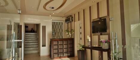 A modern hotel lobby with a reception desk, a staircase, and a flat-screen TV mounted on the wall.